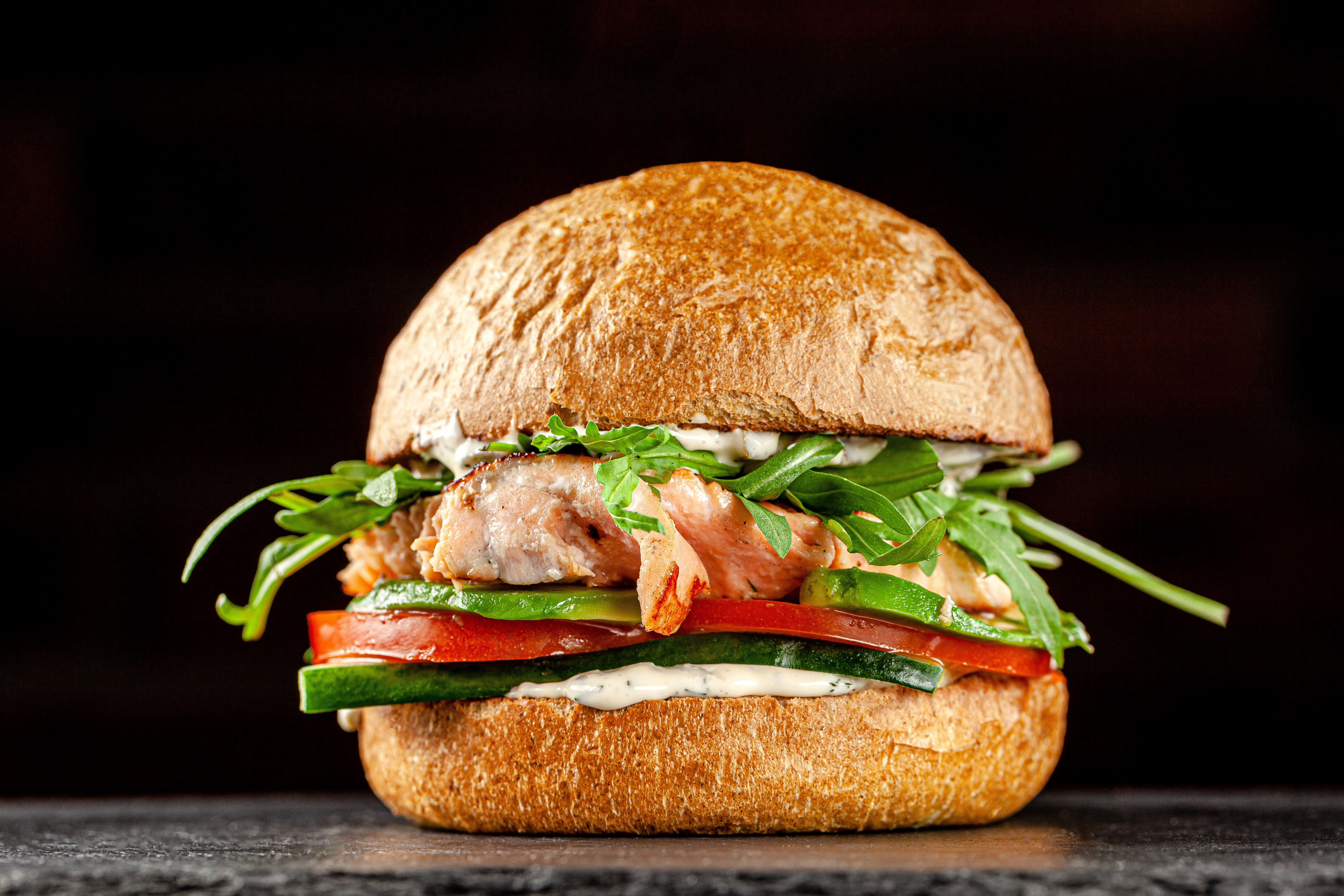 Burger with red fish salmon, arugula, cucumbers and tomatoes, white sauce. Burger on a black background. background image, copy space text